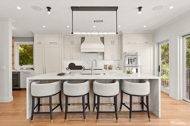 kitchen featuring premium range hood, a center island with sink, stainless steel double oven, and white cabinetry