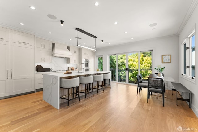 kitchen with pendant lighting, light wood-type flooring, white cabinets, custom exhaust hood, and a center island with sink