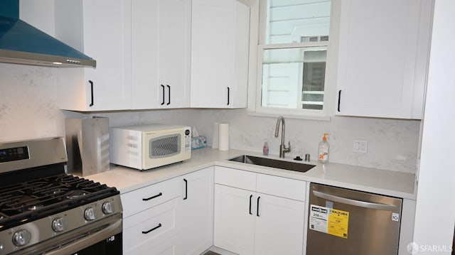 kitchen featuring appliances with stainless steel finishes, white cabinetry, a sink, light countertops, and wall chimney exhaust hood