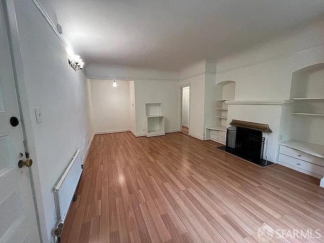 unfurnished living room featuring baseboards, a fireplace with raised hearth, radiator, wood finished floors, and built in shelves