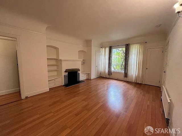 unfurnished living room with a fireplace with raised hearth, wood finished floors, baseboards, built in features, and radiator heating unit