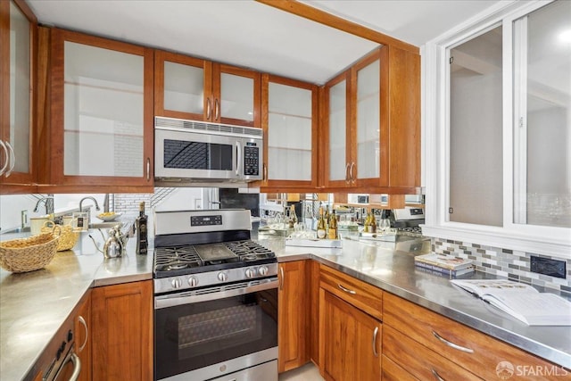 kitchen featuring backsplash, glass insert cabinets, stainless steel counters, appliances with stainless steel finishes, and brown cabinetry