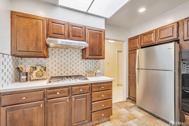 kitchen with brown cabinets, light countertops, under cabinet range hood, appliances with stainless steel finishes, and tasteful backsplash