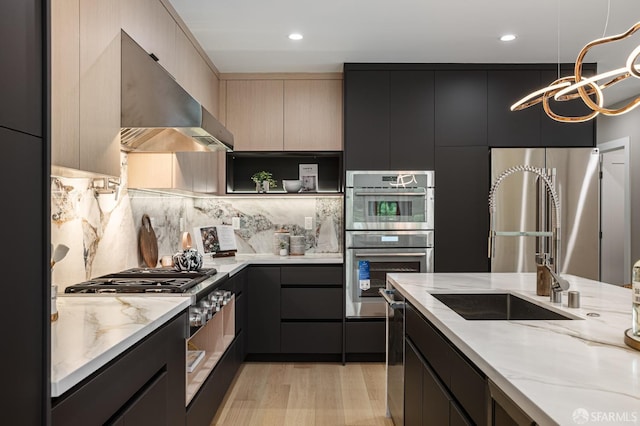 kitchen featuring wall chimney range hood, stainless steel appliances, light wood-type flooring, light stone countertops, and dark cabinetry
