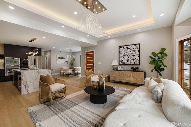 living area with a chandelier, light wood-style floors, recessed lighting, a raised ceiling, and baseboards