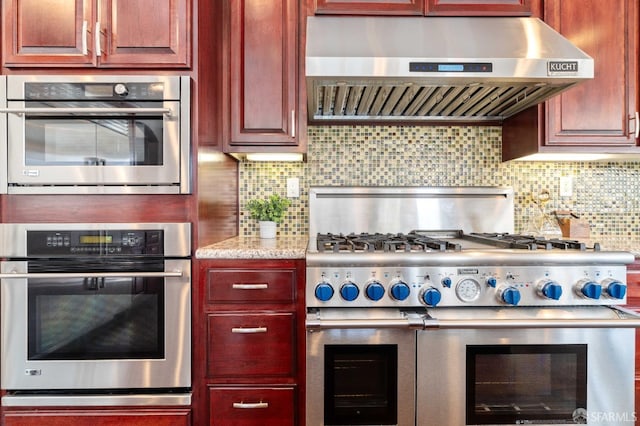 kitchen with range hood, light stone counters, reddish brown cabinets, range with two ovens, and tasteful backsplash