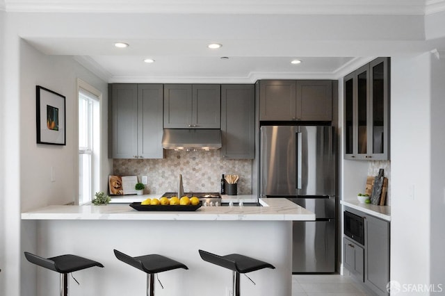 kitchen featuring freestanding refrigerator, under cabinet range hood, a breakfast bar, tasteful backsplash, and ornamental molding
