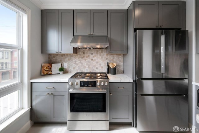 kitchen featuring appliances with stainless steel finishes, under cabinet range hood, gray cabinetry, backsplash, and light stone counters