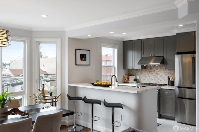 kitchen featuring freestanding refrigerator, under cabinet range hood, light countertops, plenty of natural light, and ornamental molding