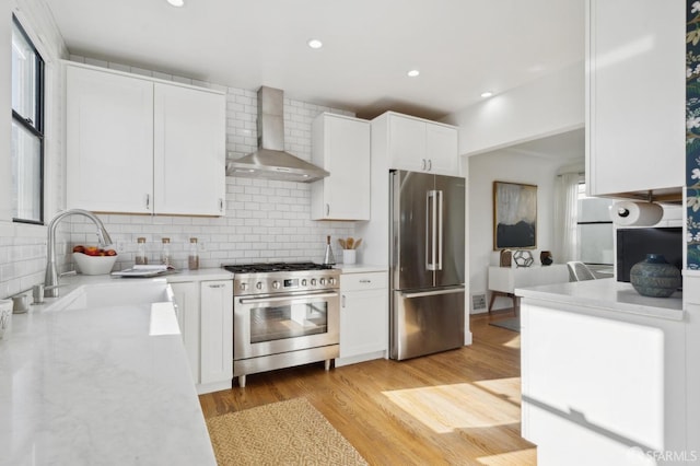 kitchen featuring light wood-style flooring, appliances with stainless steel finishes, white cabinetry, a sink, and wall chimney exhaust hood