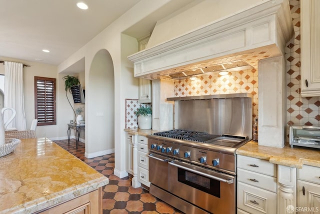 kitchen with backsplash, baseboards, premium range hood, range with two ovens, and arched walkways