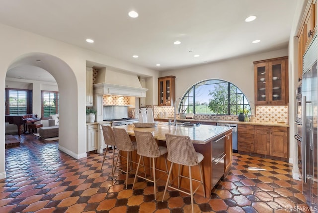 kitchen featuring a kitchen island with sink, custom range hood, plenty of natural light, arched walkways, and brown cabinetry