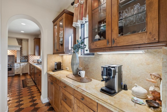 kitchen with backsplash, arched walkways, light countertops, glass insert cabinets, and custom exhaust hood