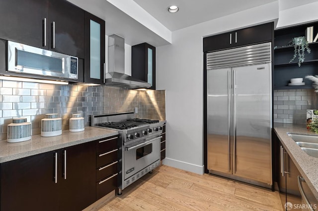 kitchen featuring premium appliances, wall chimney exhaust hood, light stone countertops, light wood-type flooring, and baseboards