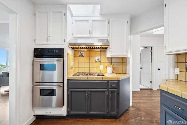 kitchen with tile countertops, stainless steel appliances, exhaust hood, and white cabinets