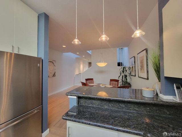 kitchen featuring hanging light fixtures, light wood-type flooring, stainless steel refrigerator, white cabinets, and dark stone countertops