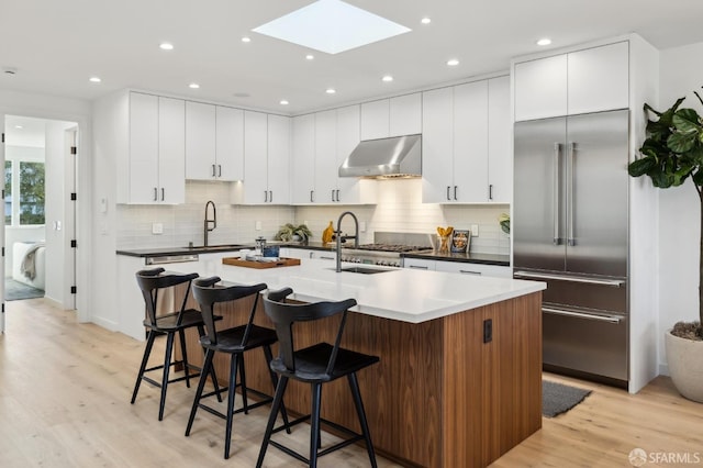 kitchen featuring built in refrigerator, light hardwood / wood-style floors, a kitchen island with sink, and range hood