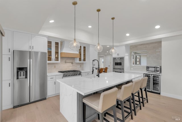 kitchen with light stone countertops, white cabinets, stainless steel appliances, a center island with sink, and custom range hood