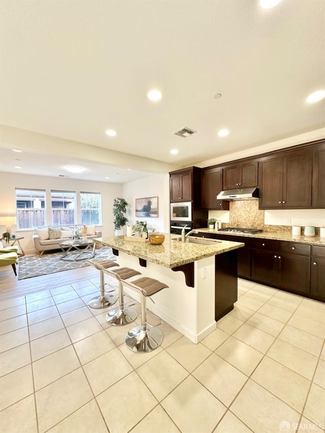 kitchen with white microwave, light tile patterned floors, a kitchen breakfast bar, a kitchen island with sink, and light stone countertops