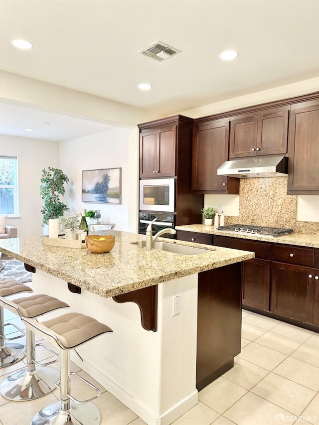 kitchen with appliances with stainless steel finishes, sink, a kitchen bar, an island with sink, and light stone counters