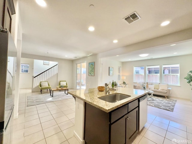 kitchen featuring a center island with sink, light stone counters, dishwasher, dark brown cabinetry, and sink