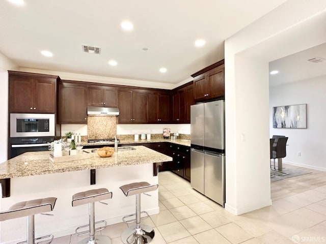 kitchen featuring light stone countertops, appliances with stainless steel finishes, an island with sink, a kitchen bar, and light tile patterned floors