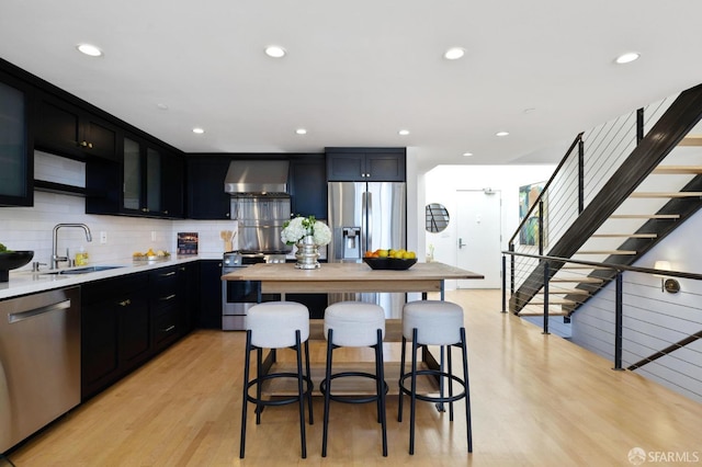 kitchen with a breakfast bar, sink, stainless steel appliances, wall chimney range hood, and light hardwood / wood-style flooring