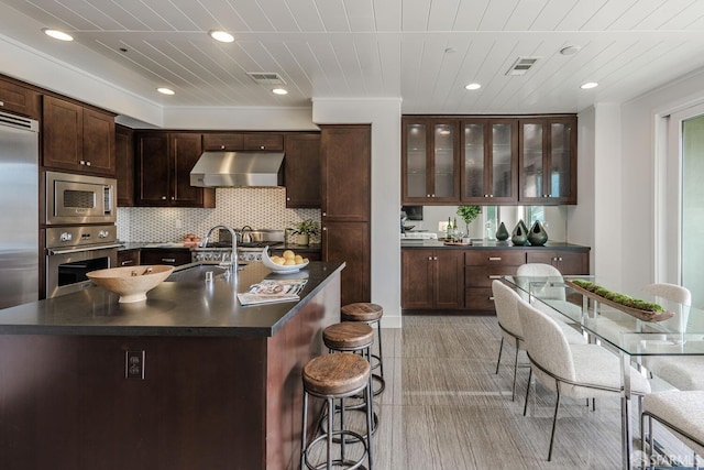 kitchen with dark countertops, visible vents, a sink, built in appliances, and under cabinet range hood