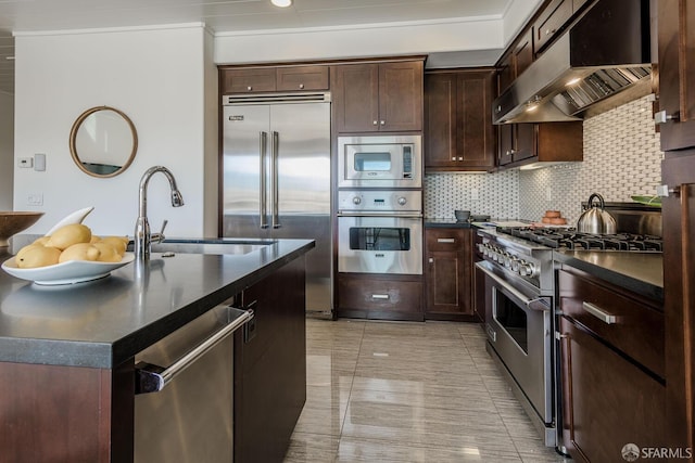 kitchen featuring tasteful backsplash, dark countertops, a sink, ventilation hood, and built in appliances