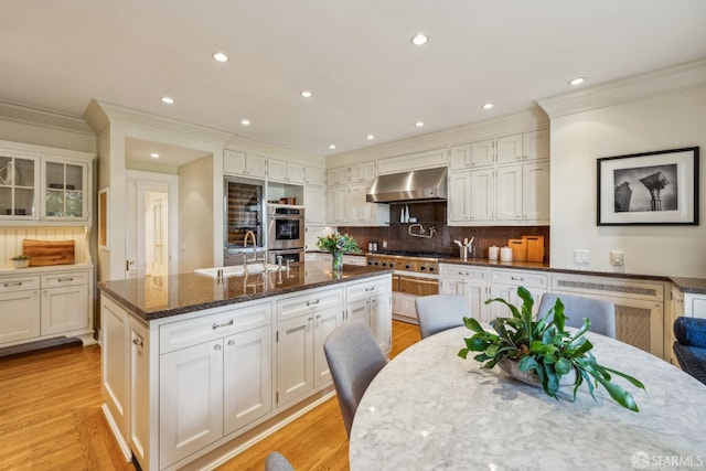 kitchen with dark stone countertops, an island with sink, a sink, light wood-style floors, and wall chimney exhaust hood