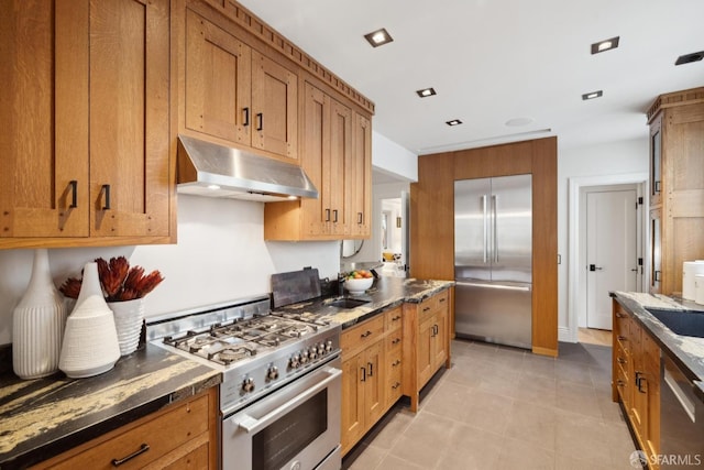 kitchen with brown cabinetry, dark stone counters, under cabinet range hood, and high quality appliances