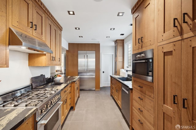 kitchen with stainless steel appliances, brown cabinetry, a sink, dark stone counters, and under cabinet range hood