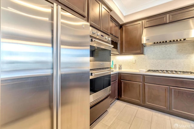 kitchen featuring appliances with stainless steel finishes, wall chimney exhaust hood, tasteful backsplash, ornamental molding, and dark brown cabinetry