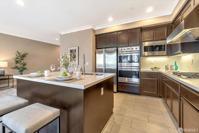 kitchen featuring appliances with stainless steel finishes, wall chimney exhaust hood, sink, an island with sink, and a breakfast bar area