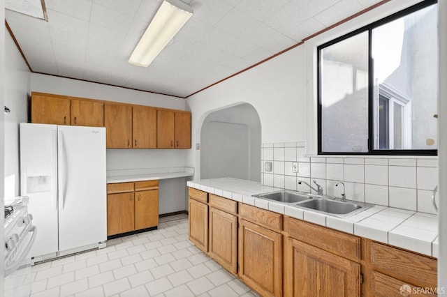 kitchen with tile countertops, white refrigerator with ice dispenser, backsplash, sink, and range