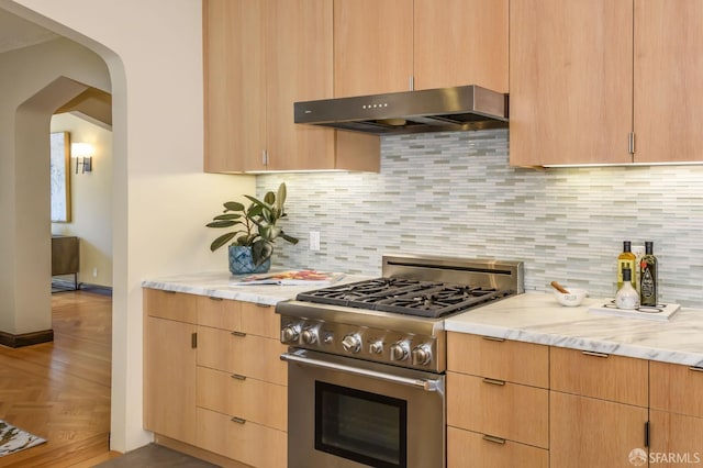 kitchen with arched walkways, light stone counters, under cabinet range hood, decorative backsplash, and stainless steel range