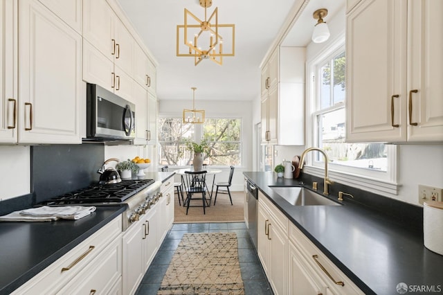 kitchen with dark countertops, appliances with stainless steel finishes, a sink, white cabinetry, and a notable chandelier