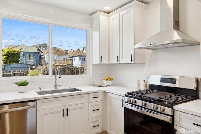 kitchen with stainless steel appliances, light countertops, wall chimney range hood, white cabinetry, and a sink