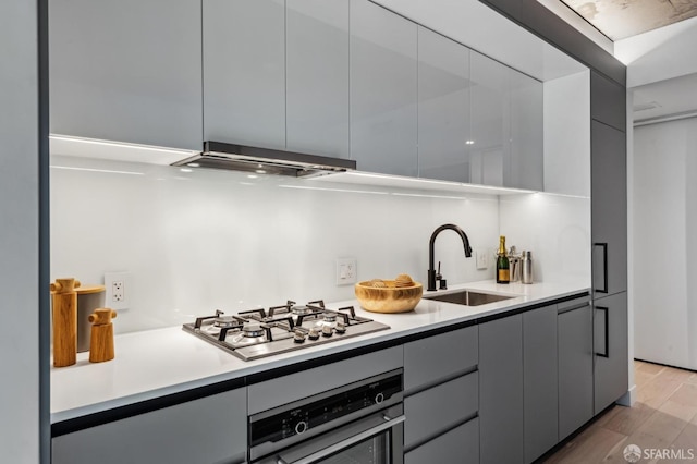 kitchen with appliances with stainless steel finishes, gray cabinetry, exhaust hood, sink, and light hardwood / wood-style flooring