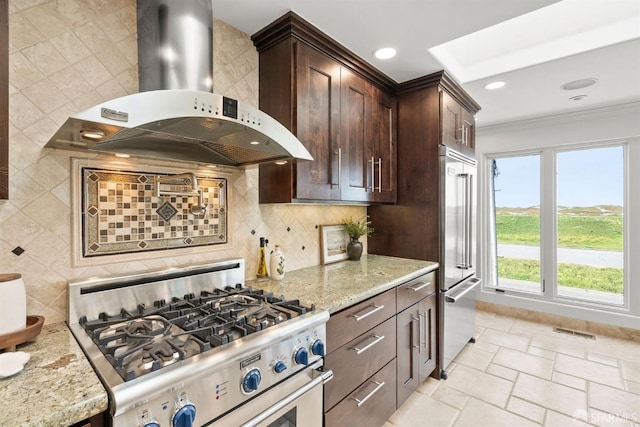 kitchen featuring light stone counters, backsplash, stainless steel appliances, wall chimney range hood, and dark brown cabinets