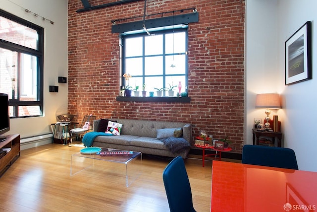 living room with baseboard heating, wood finished floors, plenty of natural light, brick wall, and a high ceiling