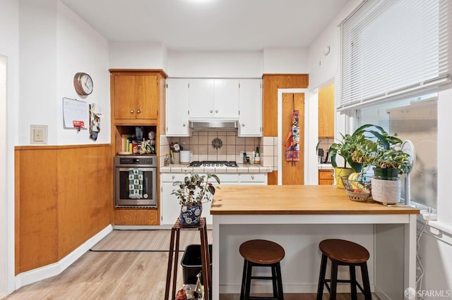 kitchen featuring a breakfast bar area, under cabinet range hood, a wainscoted wall, stainless steel appliances, and light wood finished floors