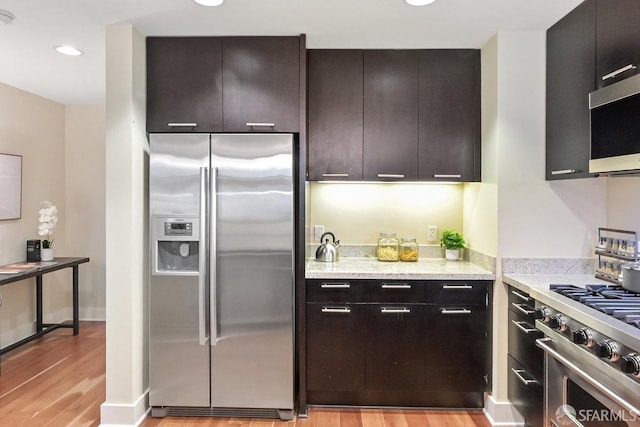 kitchen with appliances with stainless steel finishes, dark brown cabinets, light stone countertops, and light hardwood / wood-style flooring