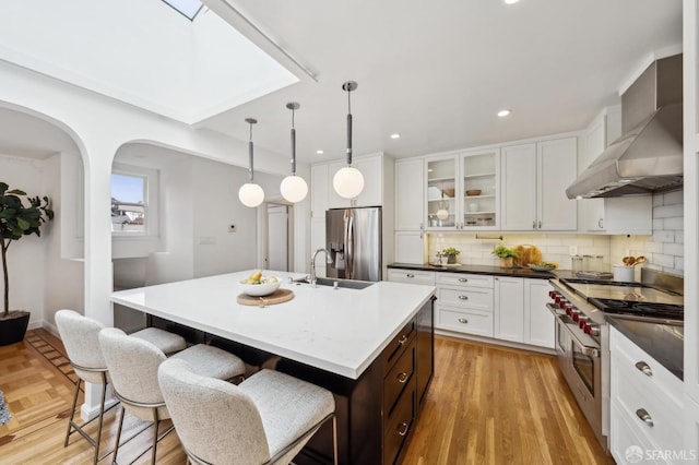 kitchen with wall chimney range hood, appliances with stainless steel finishes, a kitchen island with sink, white cabinetry, and tasteful backsplash