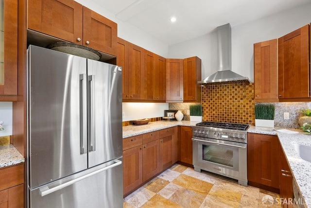 kitchen with wall chimney range hood, stainless steel appliances, brown cabinetry, and tasteful backsplash