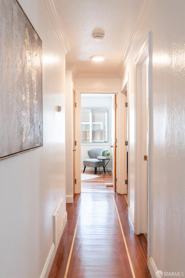 hallway with a textured ceiling, wood finished floors, visible vents, baseboards, and ornamental molding