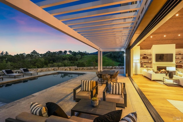 pool at dusk with a patio area and an outdoor living space with a fireplace