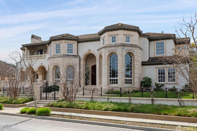 mediterranean / spanish-style home featuring a balcony, stucco siding, a fenced front yard, and a chimney