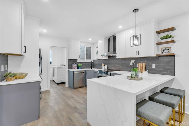 kitchen with light wood-style flooring, a peninsula, stainless steel appliances, wall chimney range hood, and washing machine and dryer