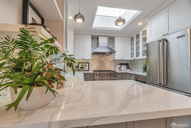 kitchen featuring white cabinets, hanging light fixtures, wall chimney range hood, light stone countertops, and glass insert cabinets